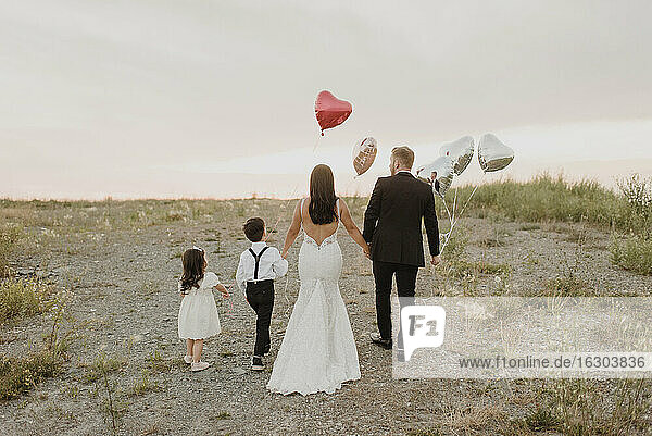 Eltern und Kinder tragen Hochzeitskleid  während sie mit herzförmigen Luftballons im Feld gegen den Himmel laufen