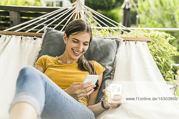Cheerful young woman holding drink using mobile phone while resting on hammock