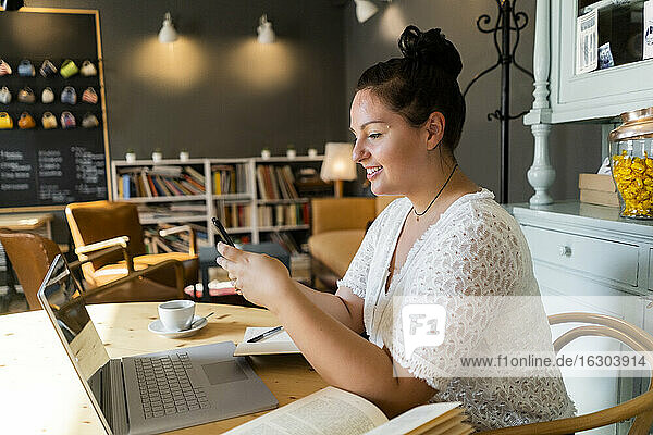 Young woman with books and laptop on table using smart phone in coffee shop