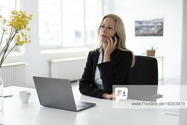 Unternehmerin mit Laptop auf dem Schreibtisch im Gespräch über ein Smartphone im Home Office