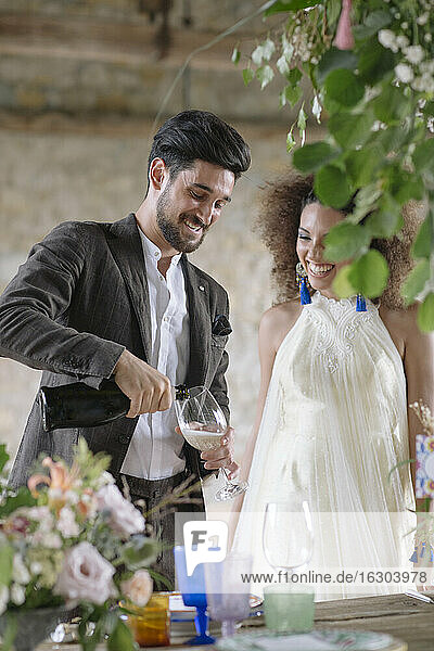 Lächelnder Bräutigam gießt Champagner in eine Flöte  während er am Tisch steht
