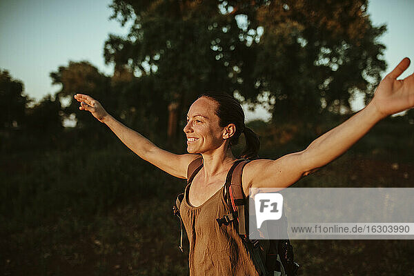 Smiling female trekker standing with arms outstretched in forest during sunset