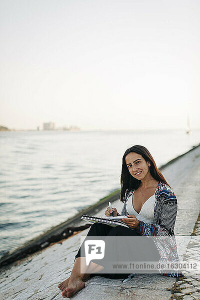 Lächelnde Frau malt in einem Buch  während sie am Flussufer sitzt