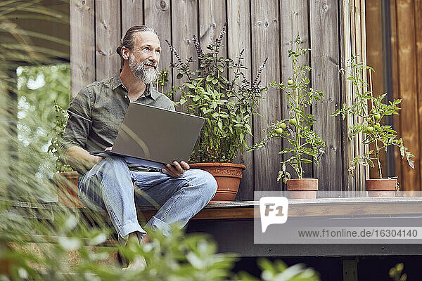 Nachdenklicher bärtiger Mann mit Laptop sitzt vor einem kleinen Haus