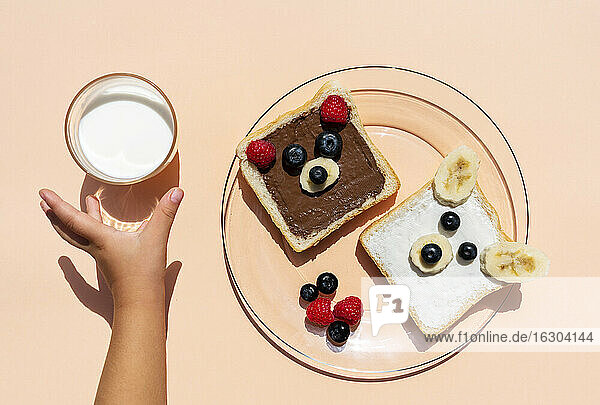 Studioaufnahme von Toasts mit Bärengesichtern aus Früchten und der Hand eines kleinen Mädchens  das nach einem Glas Milch greift