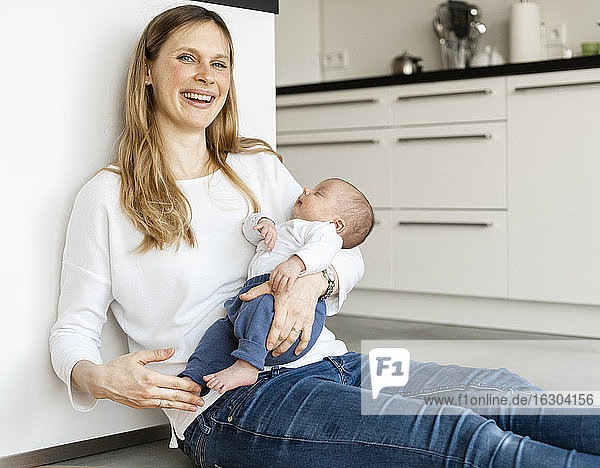 Cheerful mother holding sleeping baby boy while sitting on kitchen floor at home