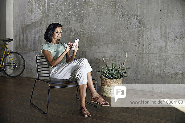Geschäftsfrau  die ein Smartphone benutzt  während sie auf einem Stuhl an der Wand im Büro sitzt