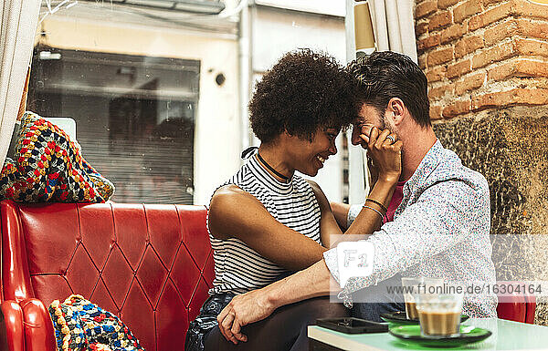 Cheerful woman pulling boyfriend's cheeks while sitting on sofa in cafe