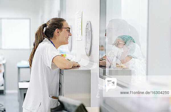 Lächelnde Ärztin im Gespräch mit einer Mitarbeiterin durch ein Fenster in einer Apotheke