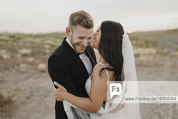 Happy groom embracing bride while standing in field against sky