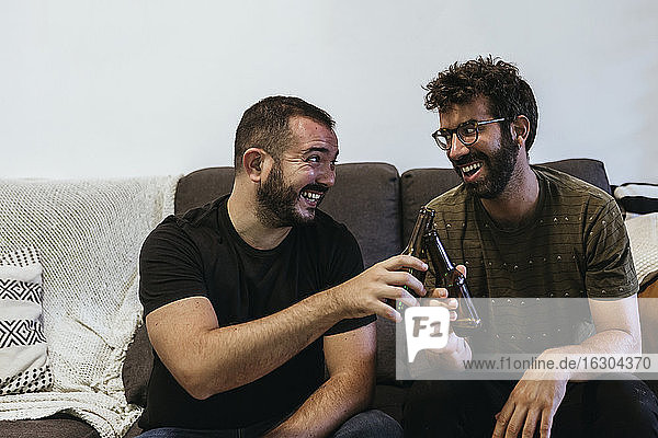 Cheerful male friends toasting beer bottles while sitting on sofa in living room