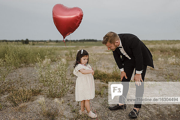 Vater spricht mit trauriger Tochter mit herzförmigem Ballon auf einem Feld gegen den klaren Himmel