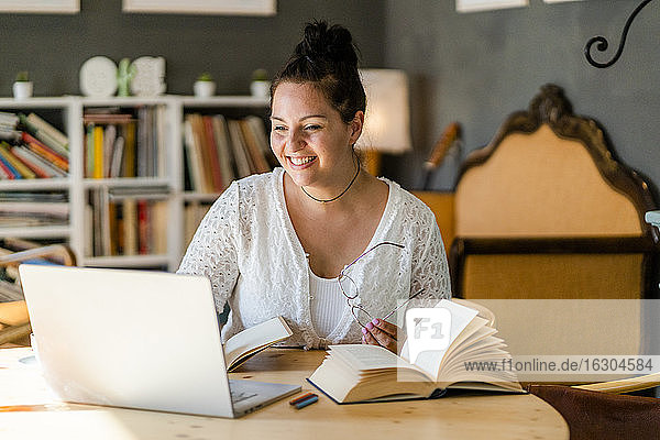 Lächelnde junge Frau  die über einem Laptop neben Büchern auf einem Tisch in einem Café studiert