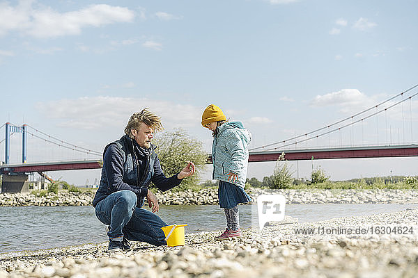 Vater hält einen Kieselstein  während seine Tochter am Flussufer steht