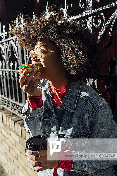 Nahaufnahme einer jungen Frau mit Afro-Haar  die an einem Zaun in der Stadt einen Donut isst