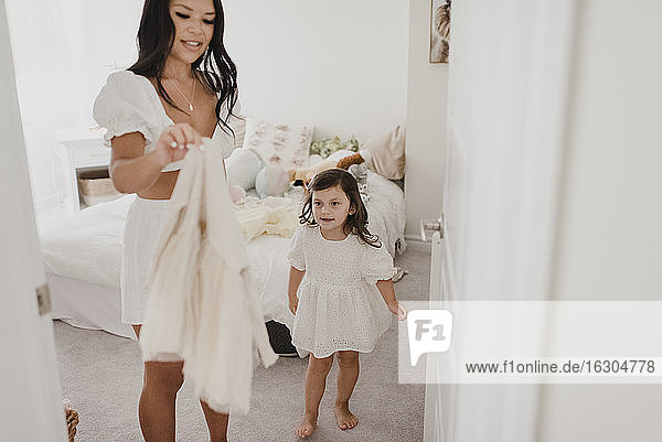 Smiling mother holding daughter's dress in bedroom
