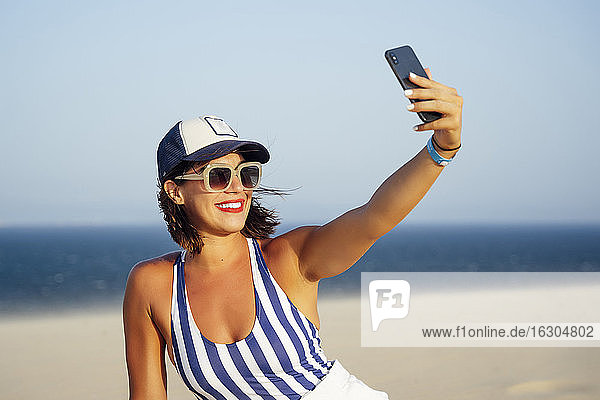 Smiling woman taking selfie while sitting at beach against clear blue sky during sunny day