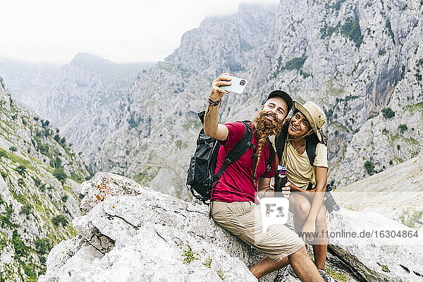 Lächelndes Paar  das ein Selfie macht  während es auf einem Felsen an der Ruta Del Cares sitzt  Asturien  Spanien