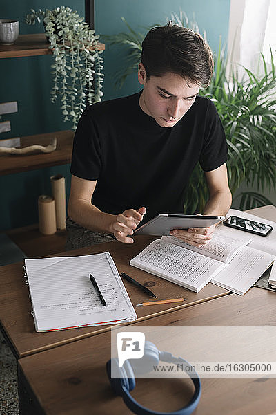 Junger Mann benutzt ein digitales Tablet  während er am Tisch sitzt und Hausaufgaben macht