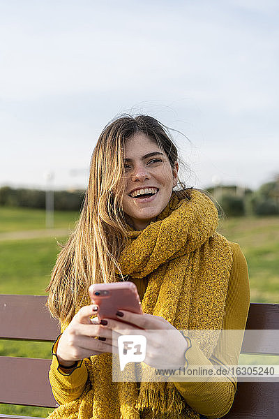 Lächelnde Frau mit Handy  die wegschaut  während sie auf einer Bank im Park sitzt