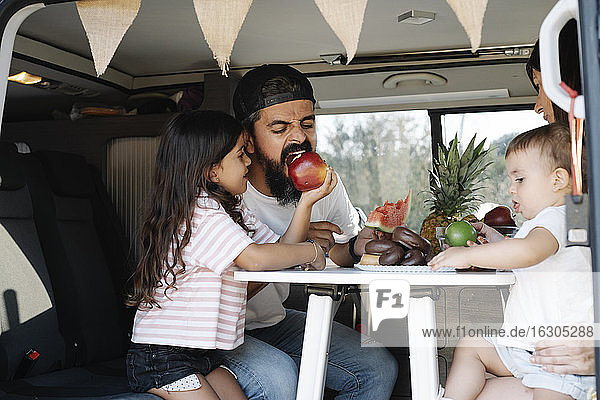 Girl feeding apple to father while sitting with mother and baby sister in van