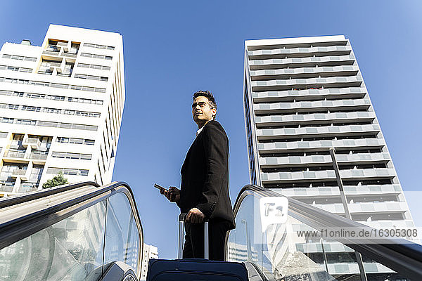 Businessman with suitcase looking away while standing on escalator against clear sky in city