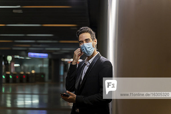 Geschäftsmann mit Maske  der sein Smartphone in der Hand hält  während er an einer Wand am Bahnhof steht