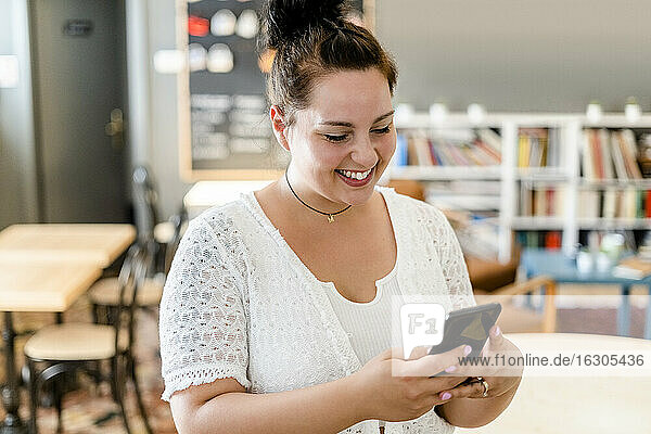 Happy young woman using smart phone while standing in coffee shop