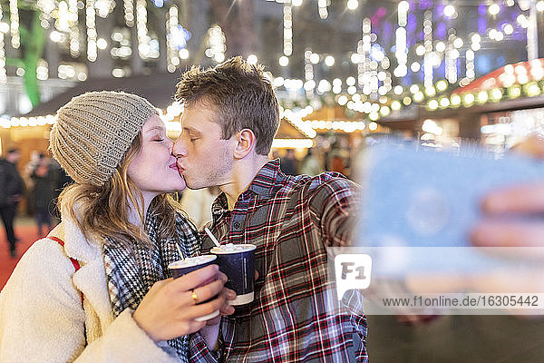 Paar hält heiße Schokolade und küsst sich  während es ein Selfie auf dem nächtlichen Weihnachtsmarkt macht