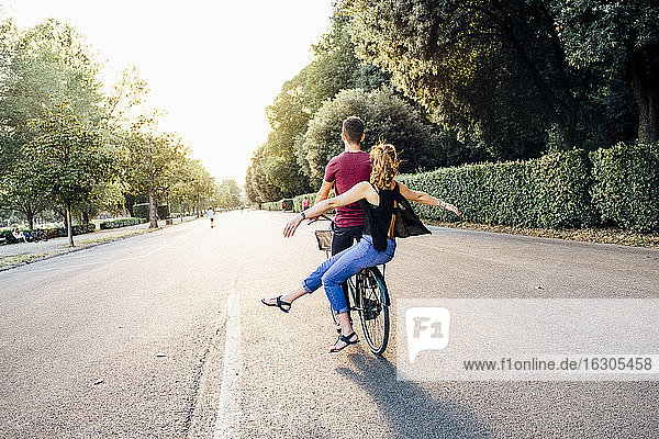 Young woman with arms outstretched sitting with boyfriend on bicycle in park