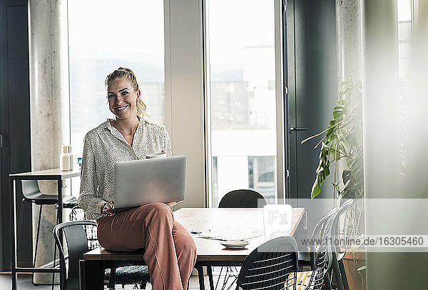 Smiling businesswoman sitting on table in office with laptop