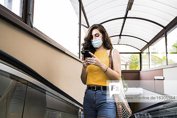 Junge Frau  die ein Mobiltelefon benutzt  während sie mit einer Netztasche auf dem Laufsteg einer U-Bahn steht