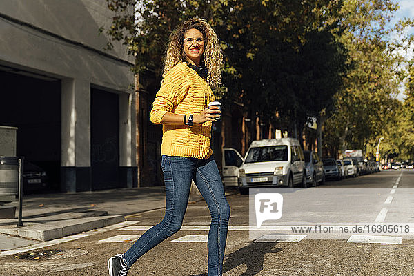 Glückliche junge Frau beim Überqueren der Straße mit einem Einwegbecher in der Stadt an einem sonnigen Tag