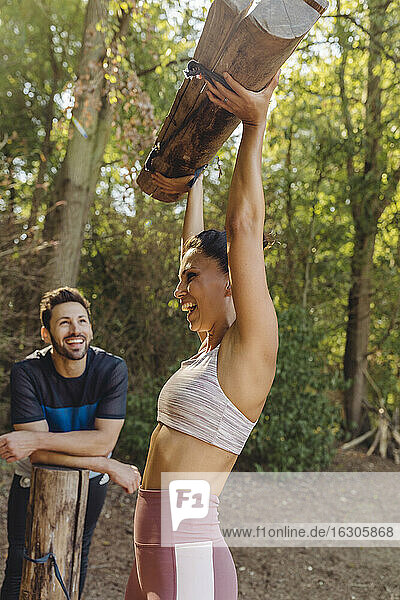 Mann beobachtet glückliche Frau  die schwere Baumstämme auf einem Fitnessparcours hochhebt