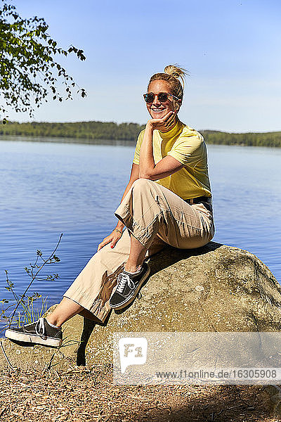 Lächelnde Frau mit Sonnenbrille sitzt auf einem Felsen am See im Tiveden-Nationalpark  Schweden