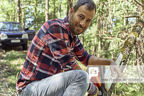 Smiling lumberjack holding chainsaw on branch in forest