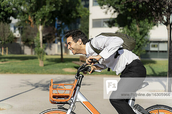 Cheerful businessman with backpack riding electric bicycle in city
