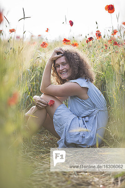 Lächelnde junge Frau mit Hand im Haar in einem Mohnfeld sitzend
