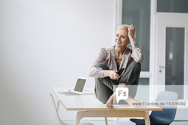 Nachdenkliche weibliche Fachkraft  die ihr Smartphone hält  während sie auf dem Schreibtisch an der Wand im Büro sitzt