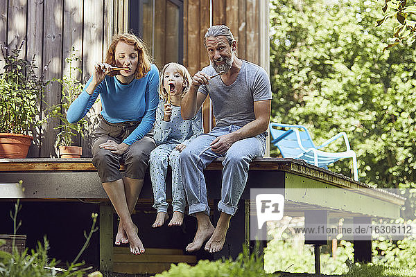 Eltern mit Tochter beim Zähneputzen  während sie vor einem kleinen Haus sitzen