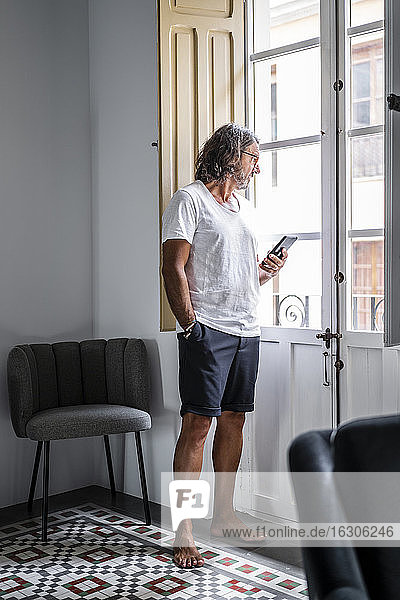 Mann mit Händen in den Taschen  der sein Smartphone hält  während er durch das Fenster seines Hauses schaut