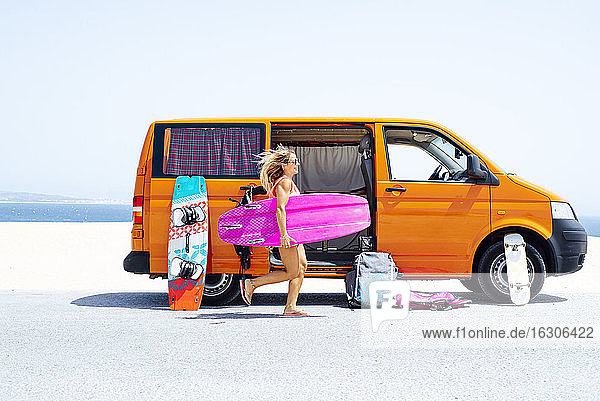 Frau mit wehenden Haaren läuft am Strand mit Surfbrett gegen Van an einem sonnigen Tag
