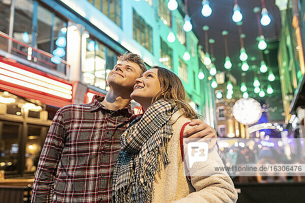 Smiling young couple looking up while standing against Christmas lights in city