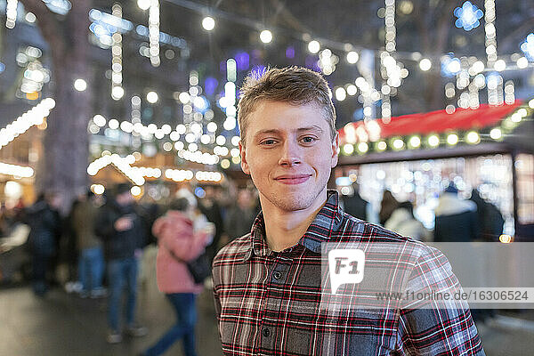 Close-up of handsome young man standing in illuminated Christmas market