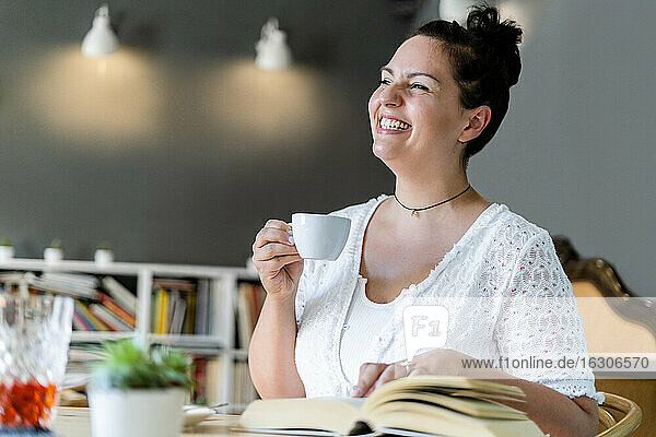 Fröhliche junge Frau schaut weg  während sie mit Kaffee und Buch in einem Café sitzt