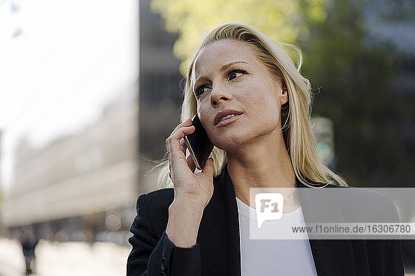 Blonde Geschäftsfrau schaut weg  während sie in der Stadt mit dem Handy telefoniert