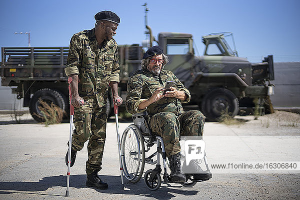 Ein verletzter Armeesoldat hält Krücken  während er mit einem behinderten Kollegen im Rollstuhl steht und auf sein Smartphone schaut.
