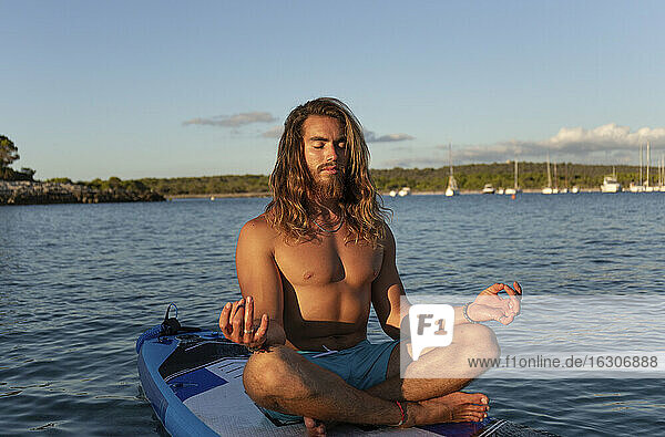 Junger Mann mit langen Haaren meditiert  während er auf einem Paddelbrett auf dem Meer gegen den Himmel bei Sonnenuntergang sitzt