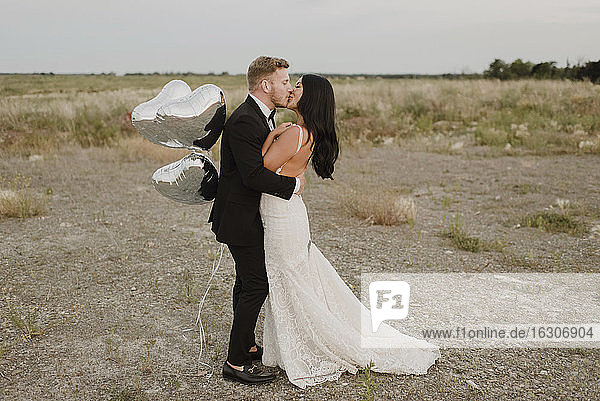 Lächelnde Bräutigam mit Herz Form Ballons küssen Braut gegen Himmel