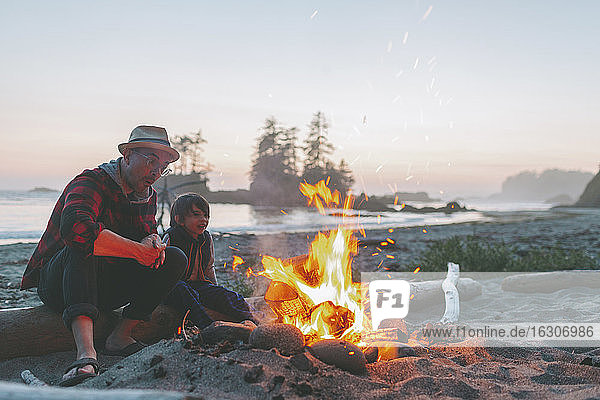 Vater und Sohn betrachten das Lagerfeuer  während sie auf Treibholz gegen den Himmel bei Sonnenuntergang sitzen